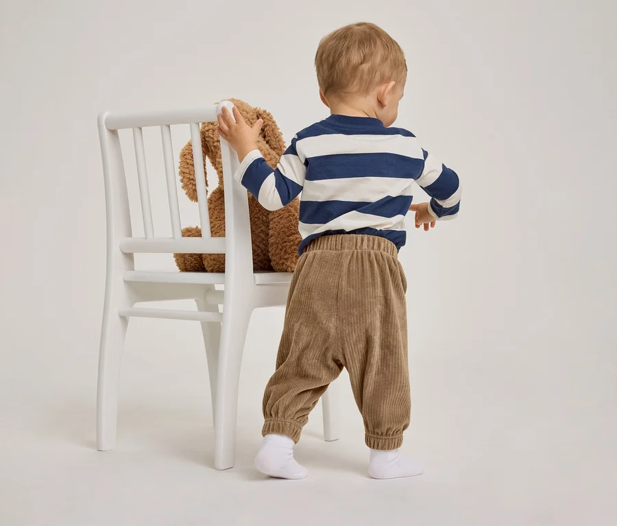 Ein Kleinkind von hinten, das sich an einem weißen Stuhl festhält. Es trägt ein blau-weiß gestreiftes Langarmshirt und eine braune Cordhose. Ein brauner Teddybär sitzt auf dem Stuhl.