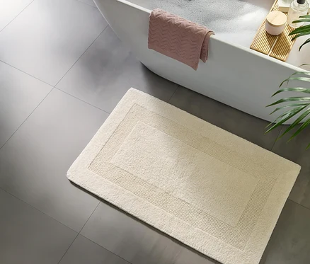 Blick von oben auf ein Badezimmer mit einer weißen Badewanne voller Schaum, einem rosa Handtuch über dem Rand und einem hölzernen Badewannenbrett. Eine cremefarbene, rechteckige Badematte mit dezentem Rand liegt auf dem grauen Fliesenboden vor der Wanne.