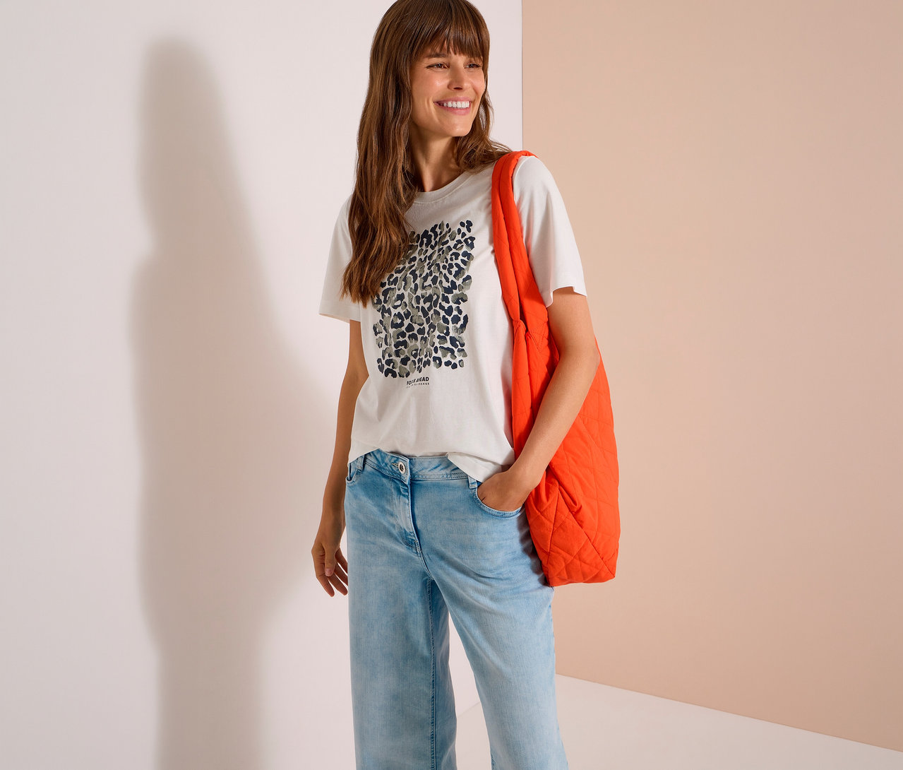 Lächelnde Frau mit langen braunen Haaren trägt ein weißes CECIL Jerseyshirt mit Leopardenmuster, hellblaue Jeans und eine orangefarbene gesteppte Tragetasche über der Schulter.