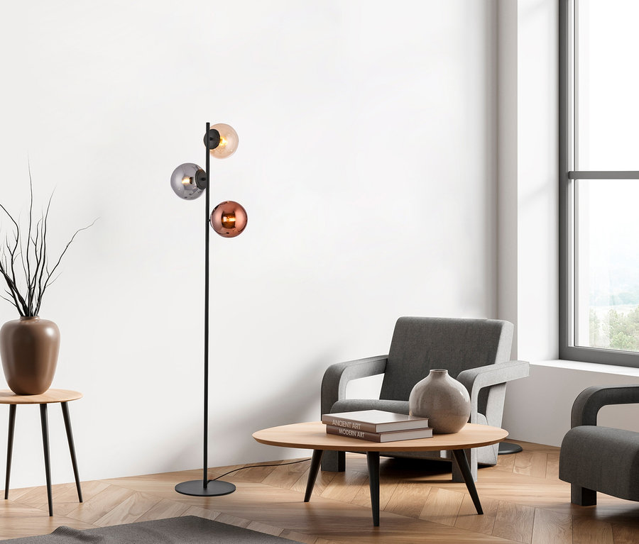 Modernes Wohnzimmer mit einer schwarzen Stehlampe, die drei kugelförmige Glasschirme in Bernstein, Grau und Kupfer hat. Zwei graue Sessel stehen um einen runden Holztisch mit einer Vase und Büchern. Links steht ein Beistelltisch mit einer Vase voller Äste. Parkettboden und ein großes Fenster sind sichtbar.