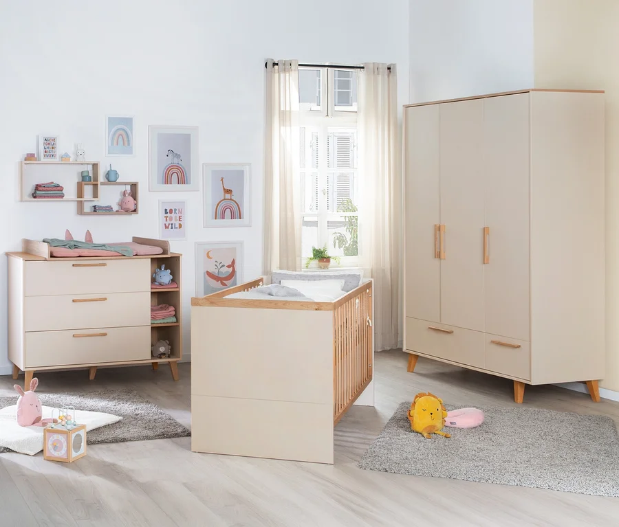 Ein helles Babyzimmer mit beiger Wickelkommode, Gitterbett und Kleiderschrank mit Holzdetails. Die Wickelkommode hat eine rosa Wickelauflage. An der Wand hängen bunte Bilder, auf dem Boden liegen graue Teppiche mit Plüschtieren und einem Holzspielzeug.