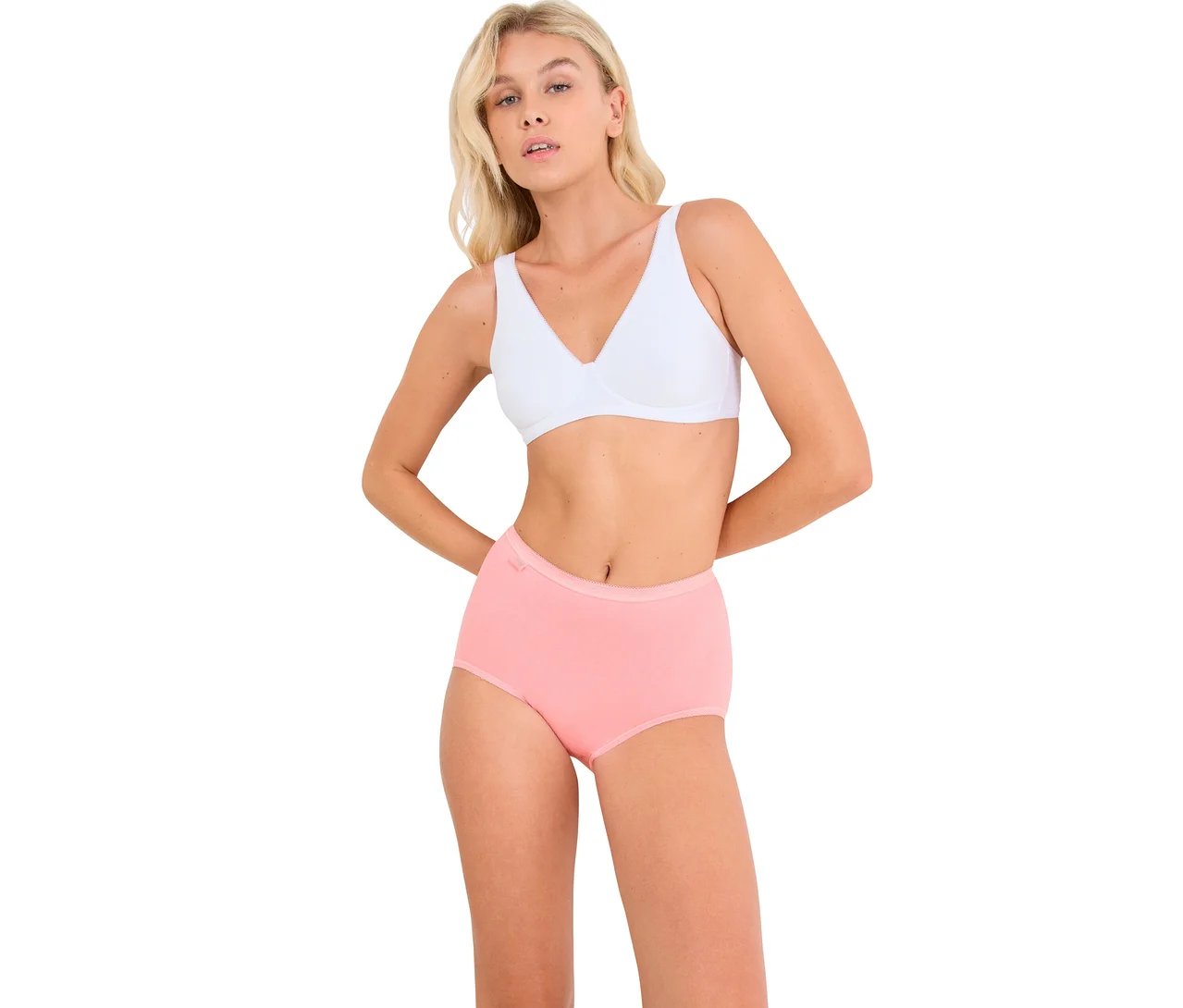 Eine blonde Frau in einem weißen BH und einem rosa Slip steht mit den Händen hinter dem Rücken und blickt nach vorne.