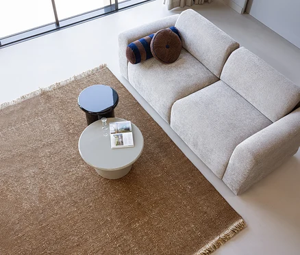 Blick von oben auf ein modernes Wohnzimmer mit einem hellgrauen Sofa, einem braunen WOOOD Teppich "Ruglio" und zwei runden Couchtischen. Der Teppich hat Fransen, der hellgraue Tisch trägt eine Zeitschrift und zwei Gläser.
