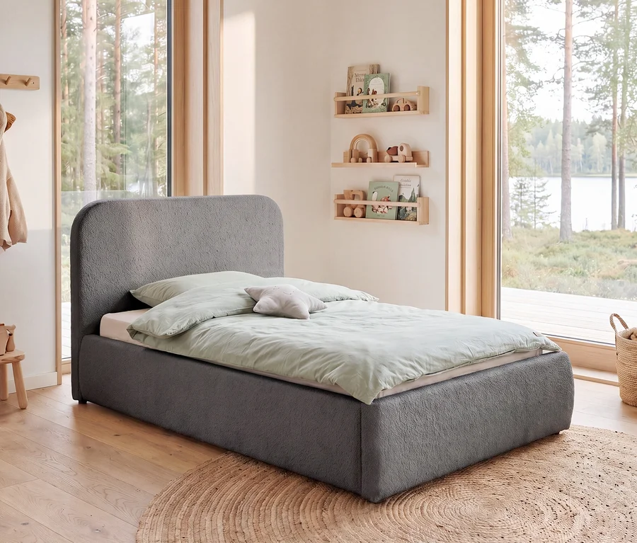 Ein graues Kinderbett mit abgerundetem Kopfteil, hellgrüner Bettwäsche und einem Sternenkissen steht in einem Zimmer mit Holzboden und Blick auf einen See. An der Wand hängen Holzregale mit Kinderbüchern und Spielzeug. Ein runder Jute-Teppich liegt auf dem Boden.