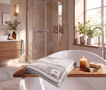 Ein hellgraues Badetuch der SCHÖNER WOHNEN-Kollektion liegt auf einem Holzbadebrett über einer Badewanne, daneben eine Kerze und Seifenstücke. Im Hintergrund sind eine Dusche und ein Waschtisch zu sehen.