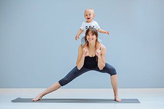 Eine glückliche, junge Mutter trägt ihr lachendes Kleinkind auf den Schultern und macht Yogaübungen auf einer Matte in Sport-Tights.