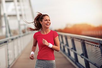 Eine junge, langhaarige Frau, die ein rotes, kurzärmeliges Sport-T-Shirt und eine graue Laufhose trägt, joggt lachend über eine Brücke