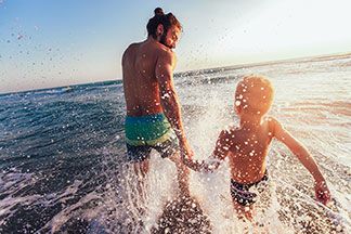 Sportbademode in guter Qualität: Ein Vater und sein kleiner Sohn, die beide Badeshorts tragen, laufen gemeinsam ins Meer und wirbeln dabei Wasser auf.