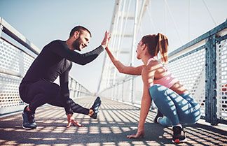 Ein Mann in schwarzer Funktionswäsche und eine junge Frau in Funktions-Sportkleidung dehnen sich in der Hocke auf einer Brücke, und klatschen sich mit der jeweils rechten Hand ab.