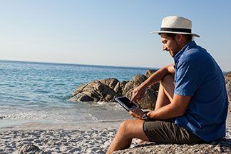 Sportbademode für den Strand: Ein junger Mann, der ein blaues Hemd, einen Hut und eine Badeshorts trägt, sitzt am Strand und schaut auf sein Tablet.