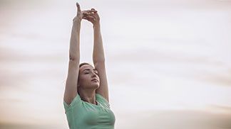 Eine junge Frau, die ein grünes Sport-T-Shirt trägt, streckt beide Arme über den Kopf nach oben und dehnt sich.