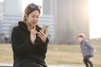 Eine junge, asiatische Frau, die mit einer dezent karierten Fleecejacke bekleidet ist, sitzt mit ihrem Smartphone vor urbaner Kulisse.