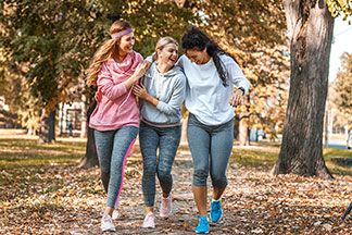Drei junge Frauen, die Sporthosen und Langarmshirts tragen, spazieren lachend durch einen herbstlichen Park.