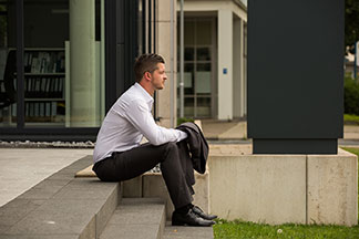Herrenhosen fürs Büro: Ein Mann sitzt in einem weißen Hemd und einer anthrazitfarbener Stoffhose auf einer Steintreppe. Das passende Jacket hält er in der Hand.
