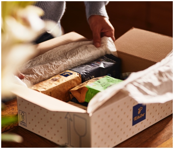 Älterer Mann packt in einer Küche ein Tchibo Paket mit Kaffee aus und hält in der einen eine Packung Feine Milde und in der anderen Hand den Bio Kaffee.