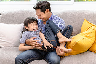 Ein Mann trägt ein blau-weiß kariertes Hemd und spielt mit seinem Sohn auf der Couch.