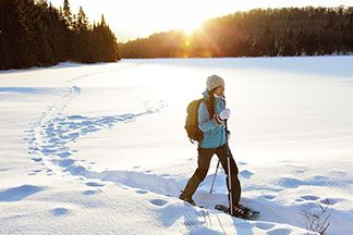 Eine junge Frau, die eine dicke, blaue Winterjacke, eine schwarze Softshellhose und eine weiße Mütze trägt, läuft mit Schneeschuhen und Stöcken durch die verschneite Winterlandschaft.
