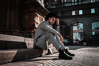Herren-Stiefel für den Businesslook: Ein junger Mann, der schwarze Boots, eine karierte Anzughose, ein kariertes Sakko sowie einen Hut trägt, sitzt im Freien auf einer Steintreppe.