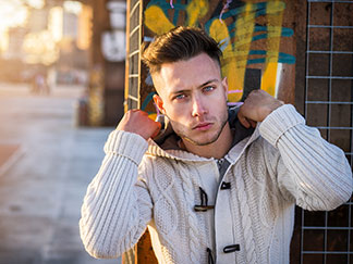 Ein junger Mann, der eine weiße rustikale Strickjacke mit Kapuze trägt, steht vor einer Wand, die mit Graffiti besprüht ist.