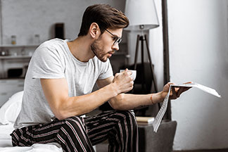 Ein Mann sitzt im Hausanzug auf dem Bett und liest sich etwas durch.