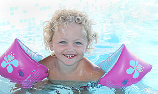 Schwimmzubehör für Kinder: Ein kleines Mädchen mit blonden Locken und rosa Schwimmflügeln schwimmt lachend im Schwimmbadbecken.