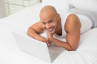 Ein Mann, der ein weißes Unterhemd und eine lange Pyjamahose trägt, liegt im Bett und arbeitet an seinem Laptop.