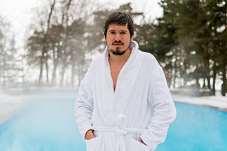 Ein bärtiger Mann in einem weißen Bademantel steht draußen vor einem dampfenden Pool und hält seine Hände in den Bademanteltaschen.