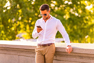 Langarm-Hemden fürs Büro: Ein Mann, der ein weißes langärmliges Hemd und eine Sonnenbrille trägt, lehnt an der Brüstung einer steinernen Brücke und bedient sein Smartphone.
