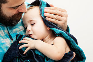 Qualitätsversprechen: Ein junger Vater hebt seine Tochter im Badezimmer bäuchlings hoch, während das Mädchen die Arme zu den Seiten hin ausstreckt.