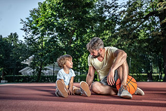 Sporthosen für die ganze Familie: Ein Vater sitzt mit seinem Sohn auf einem Sportplatz mit dem Basketball an der Seite.