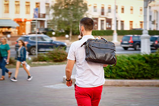 Ein junger Mann in roten Hosen und einem weißen Kurzarm-Hemd geht mit seiner Leder-Aktentasche durch die Stadt, die er über die Schulter geworfen hat.