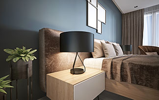 Wohlfühllicht auf dem Nachttisch: Ein hölzerner rechteckiger Nachttisch mit einer runden Lampe steht neben einem Doppelbett, auf dem eine braune Tagesdecke und beige Dekokissen liegen.