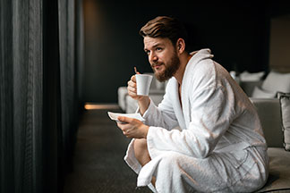 Ein junger Mann in einem weißen Bademantel trinkt, auf der Bettkante sitzend, einen Tee aus einer weißen Tasse.