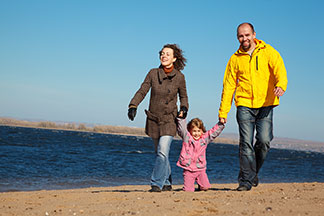 Ein Mann, eine Frau und ihre kleine Tochter, die alle Übergangsjacken tragen, gehen an einem herbstlichen Tag am Strand spazieren.