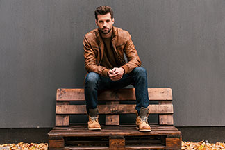 Top-Qualität für Ihre Herrenschuhe: Ein Mann, der braune Boots und eine braune Lederjacke trägt, sitzt draußen auf einer Bank aus Holzpaletten.