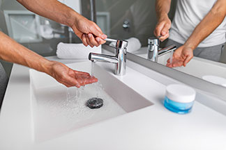 Ein Mann wäscht sich an einem weißen, eckigen Waschbecken mit einem Wasserhahn aus Metall die Hände.