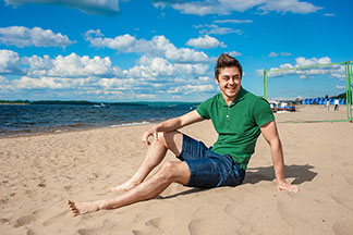 Ein junger Mann in Shorts und grünem Polohemd sitzt glücklich bei strahlendem Sonnenschein am Strand.