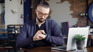 Strickjacken fürs Business: Ein junger Mann mit Brille und dunkelblauem Cardigan sitzt in einem Café vor seinem Laptop und bedient seine Smartwatch.