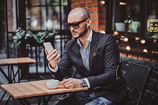 Qualität für Ihre Herrenuhr: Ein junger Mann im Anzug sitzt im Außenbereich eines Cafés und schaut auf sein Handy. Am Arm trägt er eine Herrenuhr.