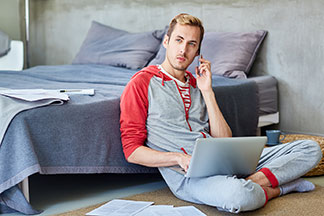 Ein junger Mann sitzt im Hausanzug vor seinem Bett und hat seinem Laptop im Schoß.