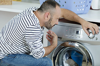 Ein Mann, der eine Jeans und einen weiß-schwarz gestreiften Pullover trägt, kniet vor der Waschmaschine, um seine Jogginghose zu waschen, und stellt das Programm ein.