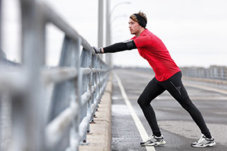 Ein junger, sportlicher Mann in einem langärmligen Funktionsunterhemd, über dem er ein rotes Shirt trägt, macht auf einer Brücke Dehnübungen während des Lauftrainings.