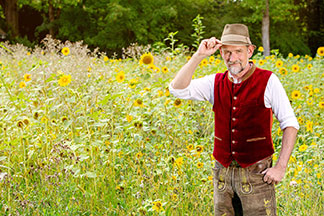Traditionelle Herrenwesten: Ein Mann steht mit Lederhose und roter Tachtenweste vor einem Sonnenblumenfeld und fasst sich an seinen Hut.