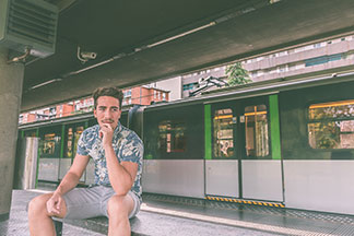 Ein junger Mann in einem Kurzarmhemd mit floralem Muster und kurzen Chinos sitzt an einer S-Bahn-Haltestelle auf einer Bank.