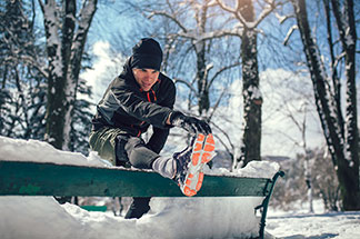 Ein junger Mann beim Joggen, der gerade in winterlicher, verschneiter Umgebung Stretchübungen an einer Bank macht.