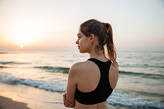 Rückansicht einer jungen Frau im schwarzen Sport-BH, die am Meer steht und den Sonnenuntergang beobachtet.
