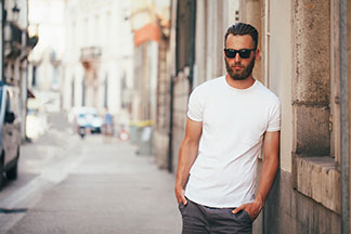 Basics für den Kleiderschrank: Ein bärtiger junger Mann mit Sonnenbrille und einem weißen Basic-Shirt steht lässig an die Mauer eines Hauses gelehnt.