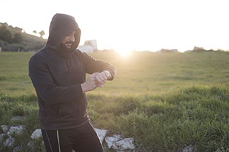 Sweatshirts für den Sport: Ein Mann, der ein schwarzes Kapuzen-Sweatshirt sowie Jogginghosen trägt, steht im Freien, hat die Kapuze aufgezogen und schaut auf seine Uhr.