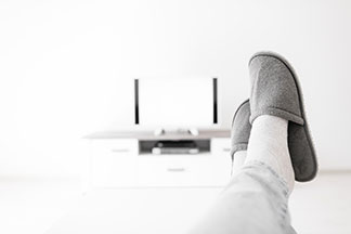 Herrenschuhe für daheim: Nahaufnahme von den Beinen eines Mannes, die ausgestreckt auf einem Tisch abgelegt sind. An den Füßen trägt der Herr Pantoffeln.