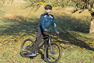 Ein bärtiger Mountainbiker mit Funktions-Jogginghose, Shirt, Helm und Sonnenbrille steht an einem sonnigen Tag an einem Hang und genießt seine Tour.